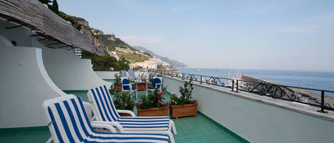 A terrace with two striped lounge chairs, a table, and potted plants. The terrace overlooks a coastal area with buildings and a clear sky.