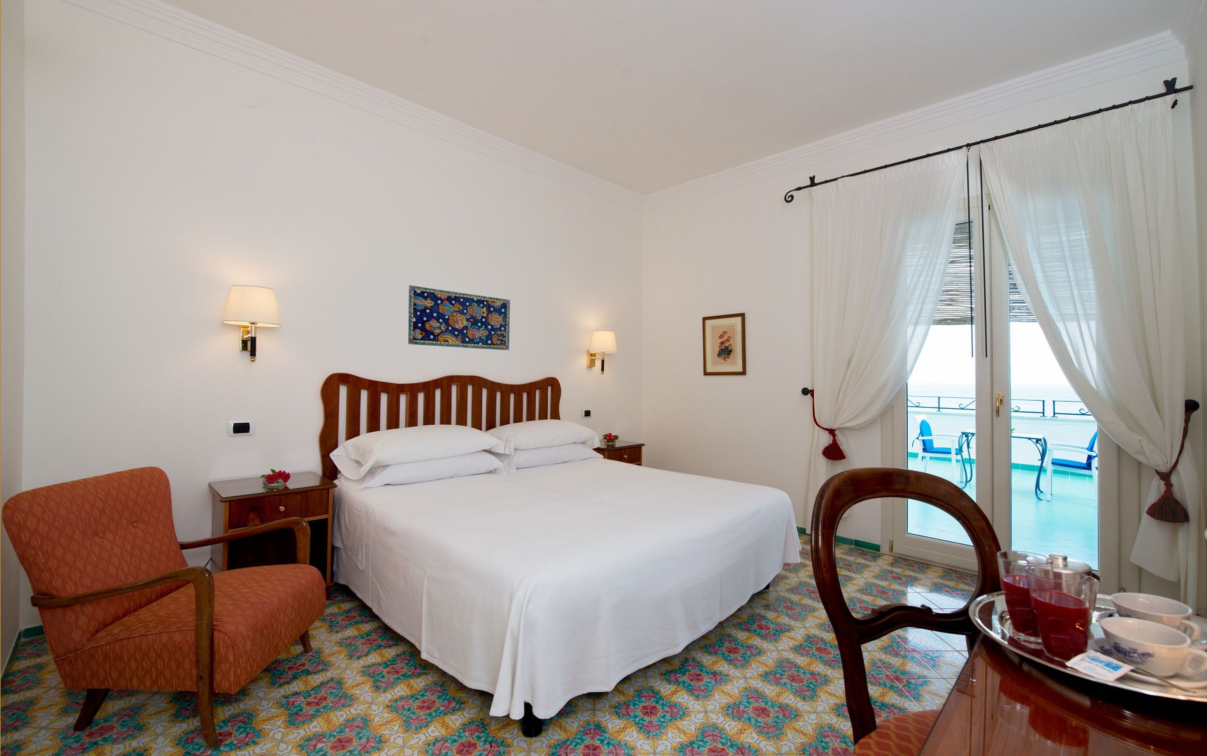 A hotel room with a bed, two chairs, a small table with a tray of glasses and cups, a nightstand, a wall-mounted lamp, and a window with curtains.