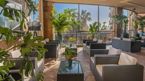 Un vaste espace de détente extérieur, aménagé avec des meubles en osier et des plantes en pot, et de grandes fenêtres offrant une vue sur les palmiers et les bâtiments.