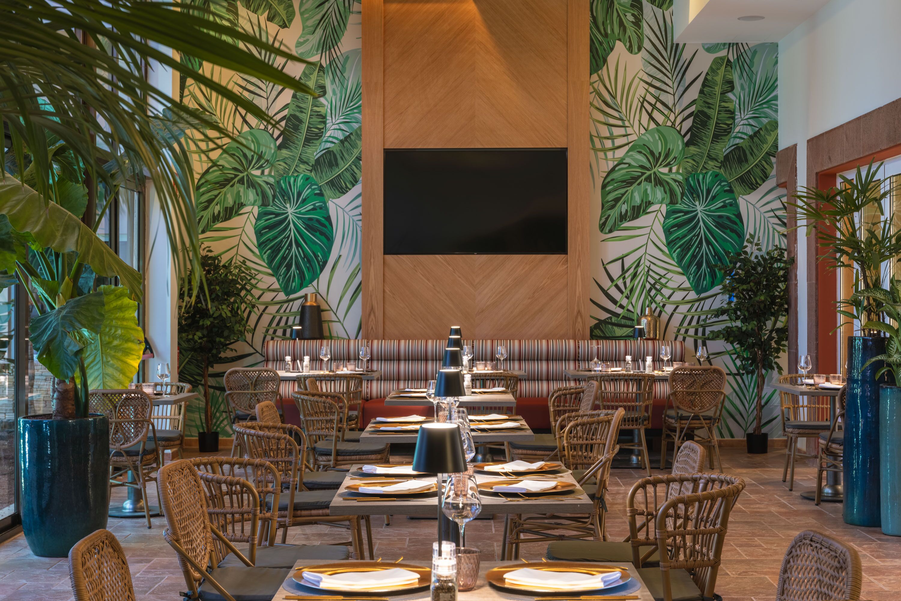 Een restaurant met een muurschildering van tropische bladeren, rieten meubels en een flatscreen televisie.