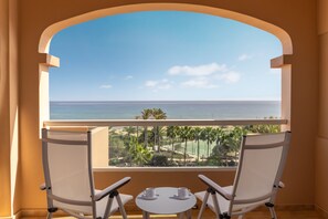 Un balcón con dos sillas blancas y una mesita, con vista a una playa y el océano.