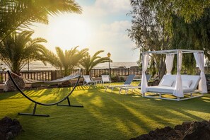 Una hamaca, sillones de descanso y una cama con dosel en un jardín con vista al océano.