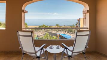A balcony with two white chairs and a small table, overlooking a beach and a pool.
