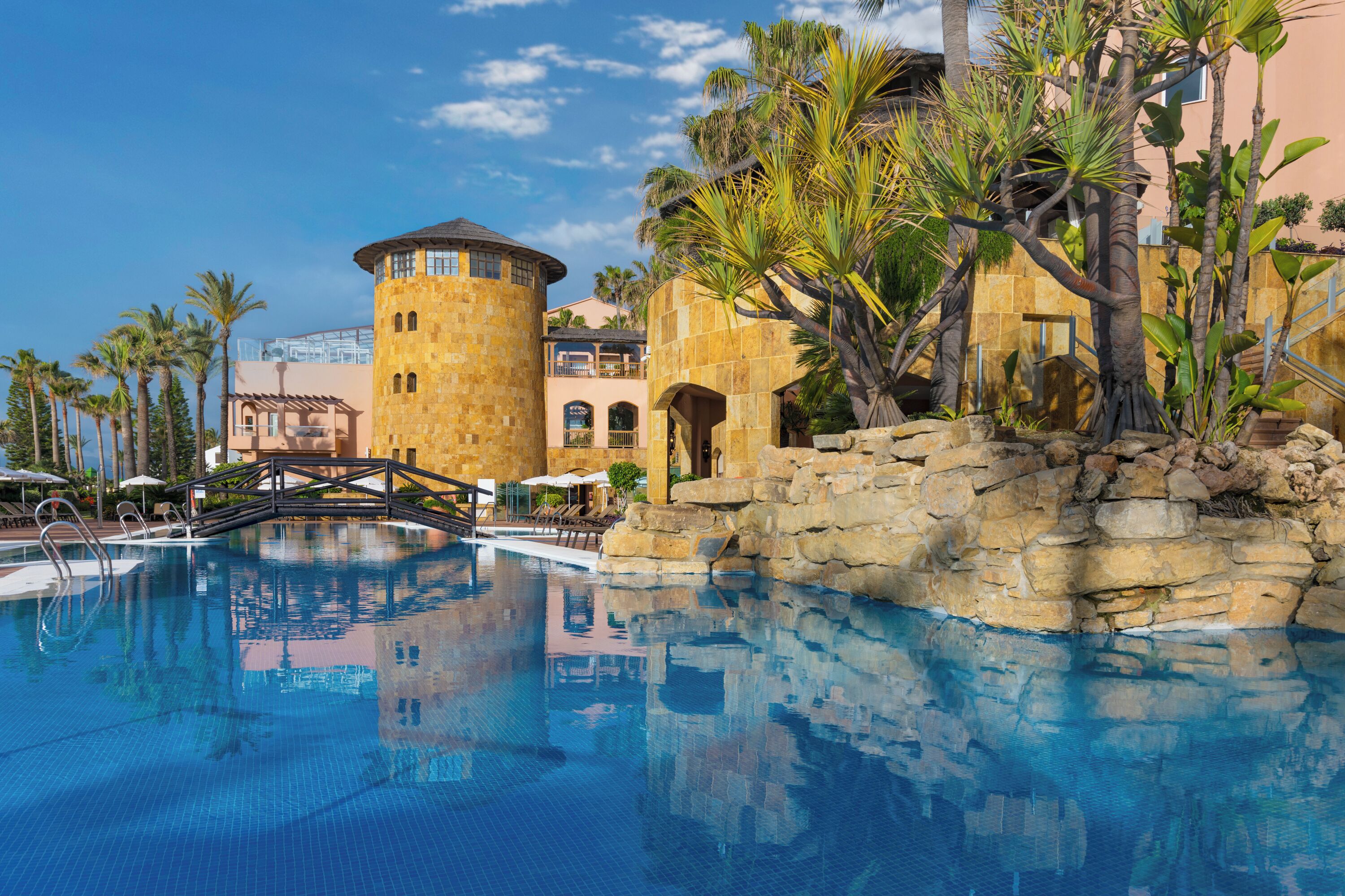 A pool area with a stone bridge, palm trees, and a building with arched windows.