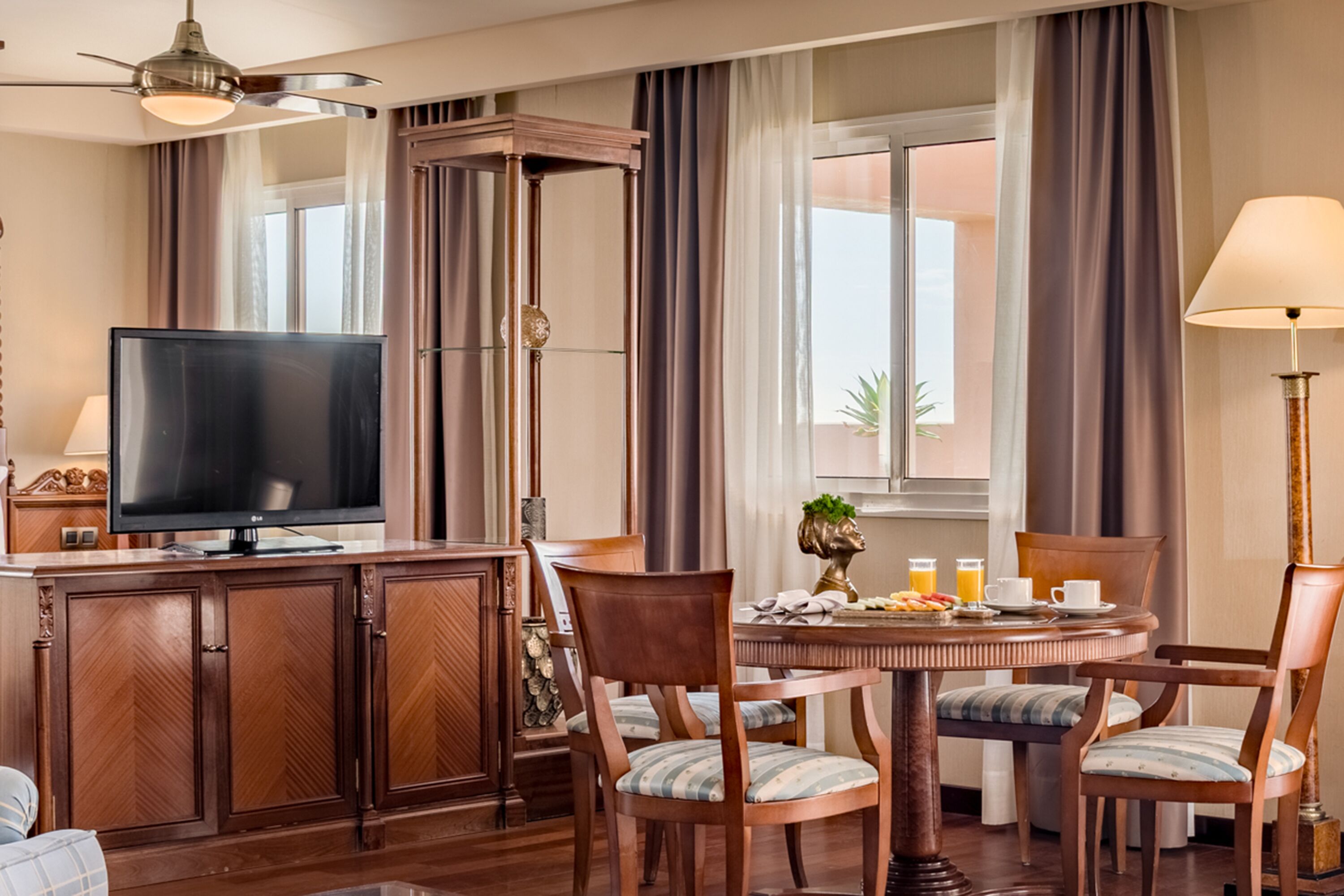 A hotel room with a dining area featuring a round table, wooden chairs, a television, and a lamp.