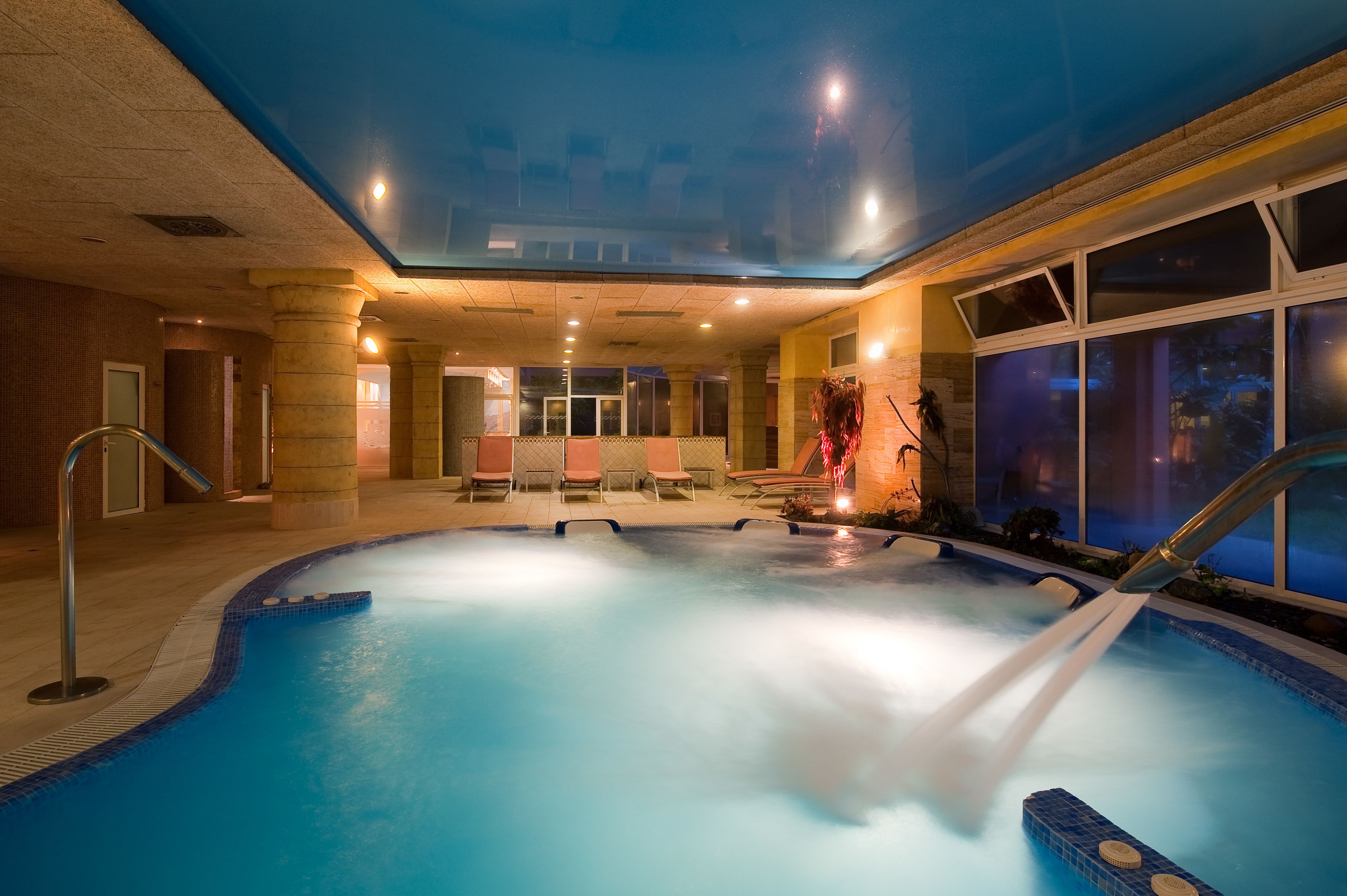 An indoor swimming pool with a hot tub, lounge chairs, and a glass ceiling.