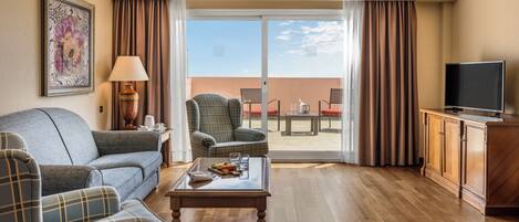A living room with a sofa, armchair, coffee table, and a television. There is a balcony with a table and chairs.