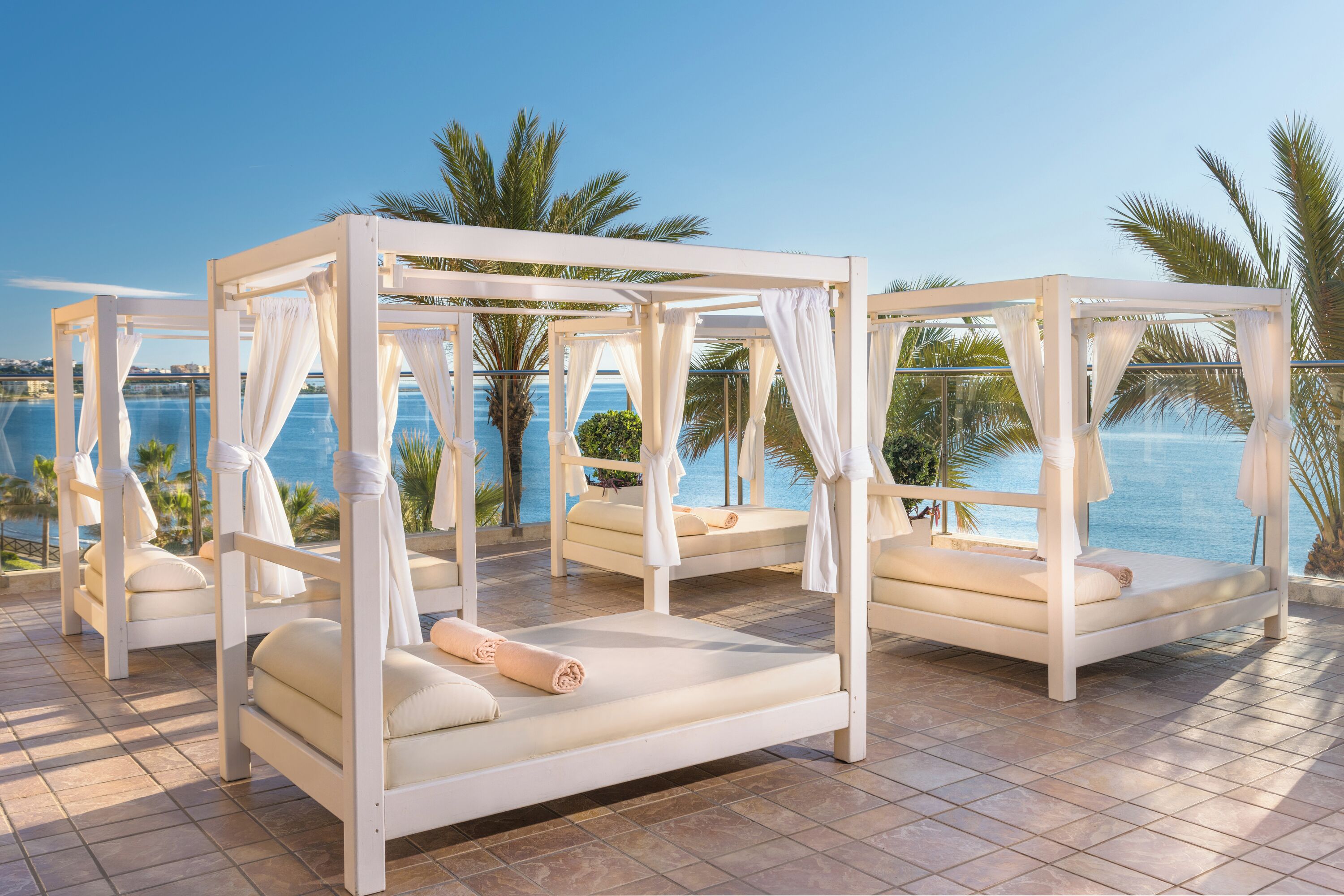 A set of four white four-poster beds with white linens, arranged on a tiled patio with a view of the sea and palm trees.