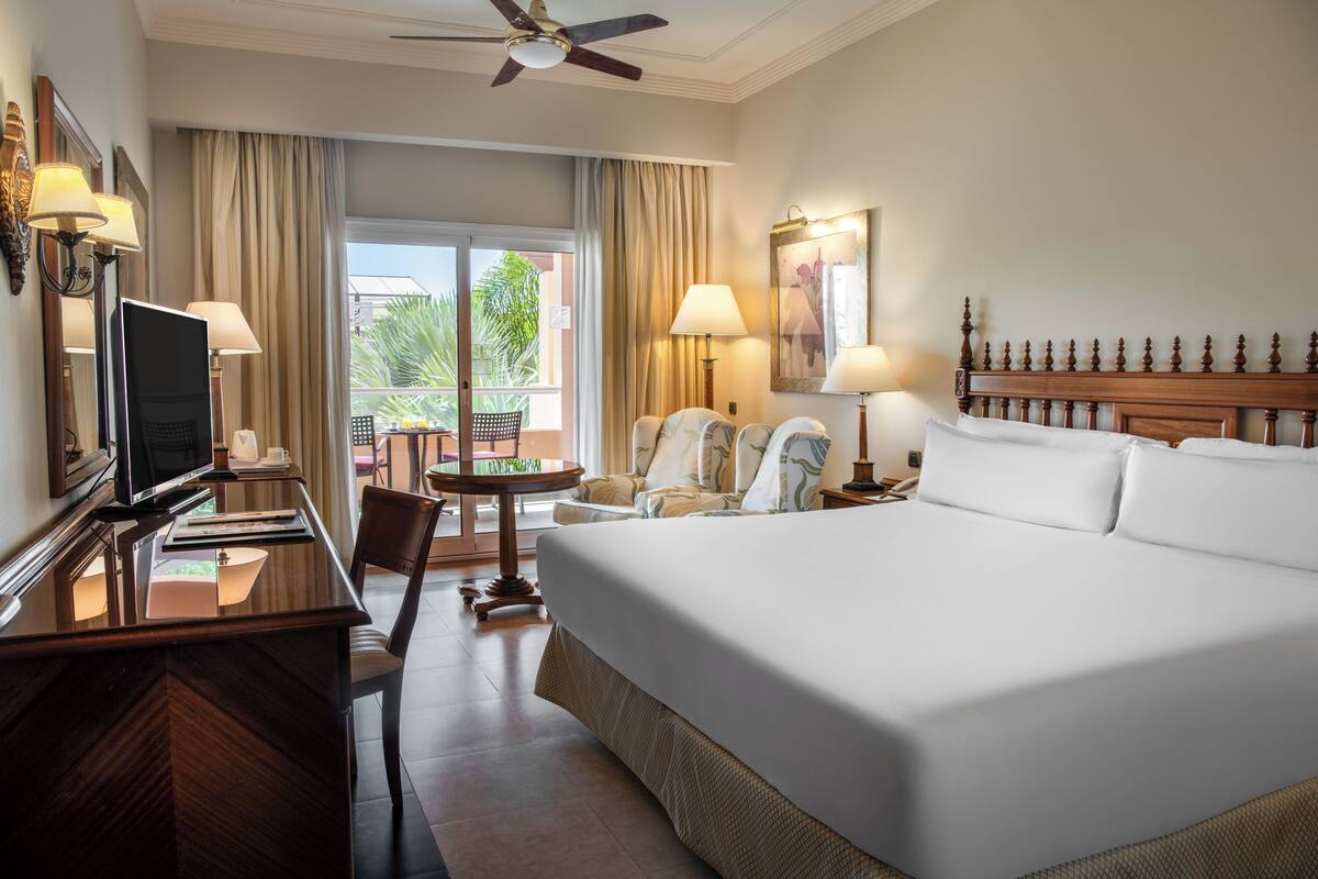 Une chambre d’hôtel avec un grand lit, un bureau avec une télévision, une chaise, une table et un balcon avec des rideaux.