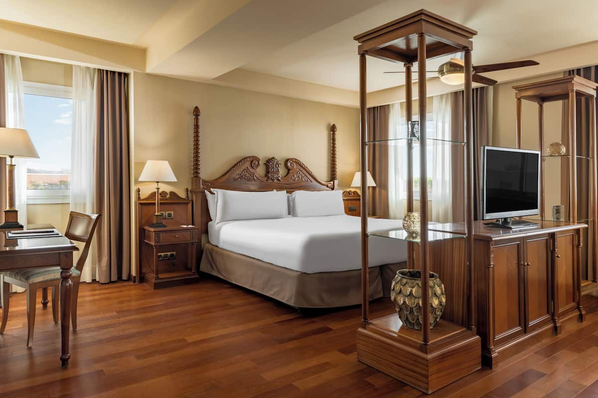 Une chambre d’hôtel avec un grand lit, une tête de lit en bois, une télévision, un bureau avec une lampe et un vase posé sur un piédestal.