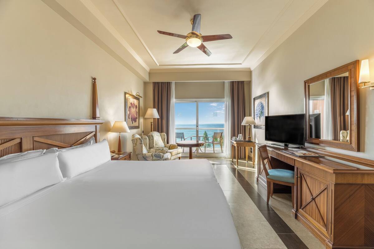 Une chambre d’hôtel avec un grand lit, un bureau avec une télévision, une chaise, une petite table et une vue sur l’océan par une grande fenêtre.
