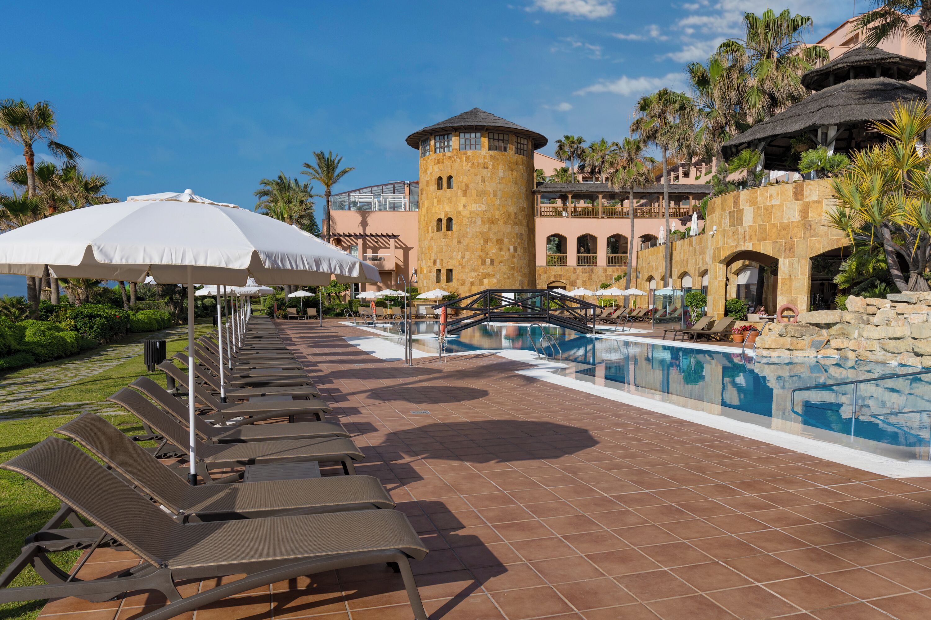 Zona de piscina en un complejo turístico, con sillas de descanso, sombrillas y una piscina central.
