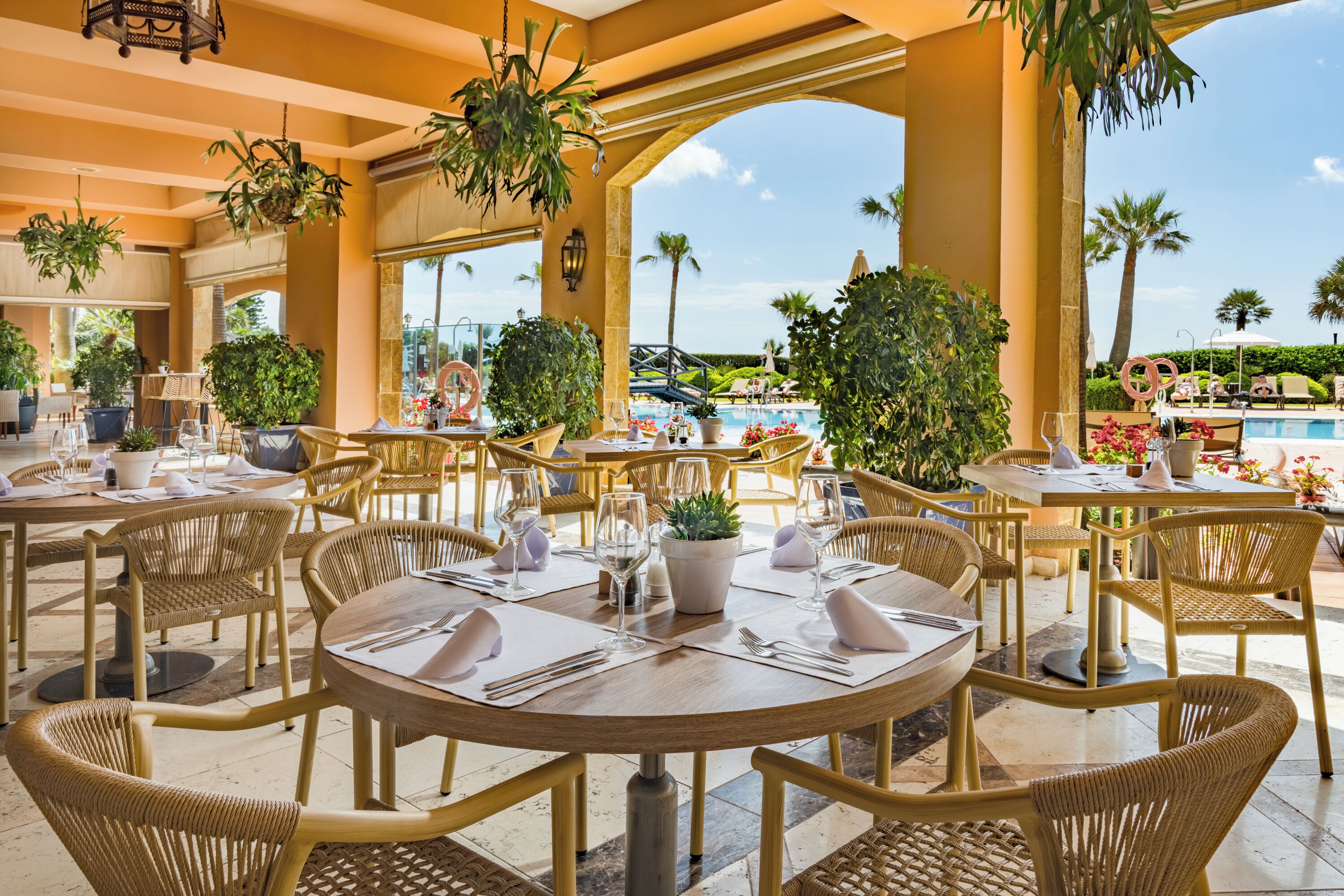 A restaurant with outdoor seating, tables set with white napkins and cutlery, surrounded by potted plants and overlooking a pool area.
