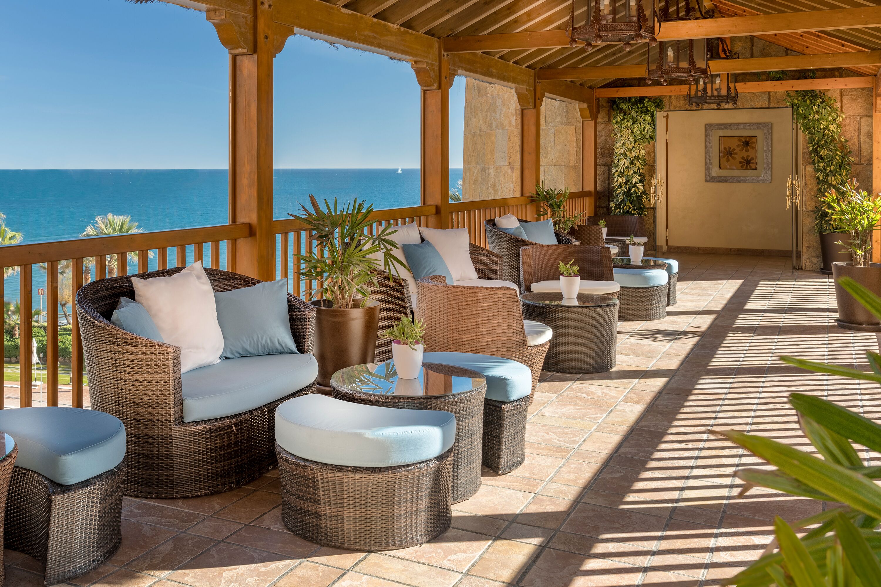 A covered outdoor seating area with wicker furniture and potted plants, overlooking the sea.