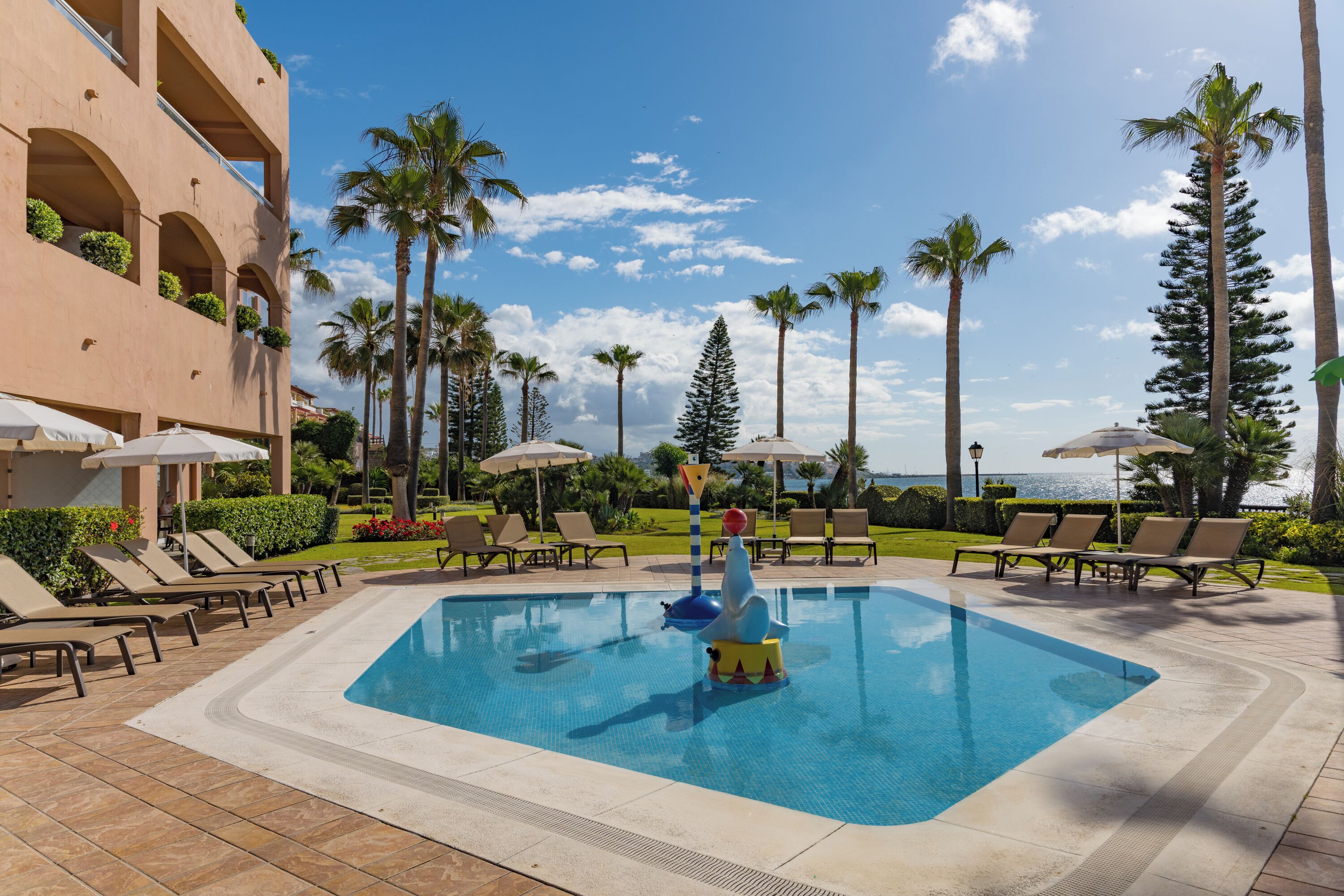 A swimming pool with lounge chairs and umbrellas, surrounded by palm trees and a building with balconies.