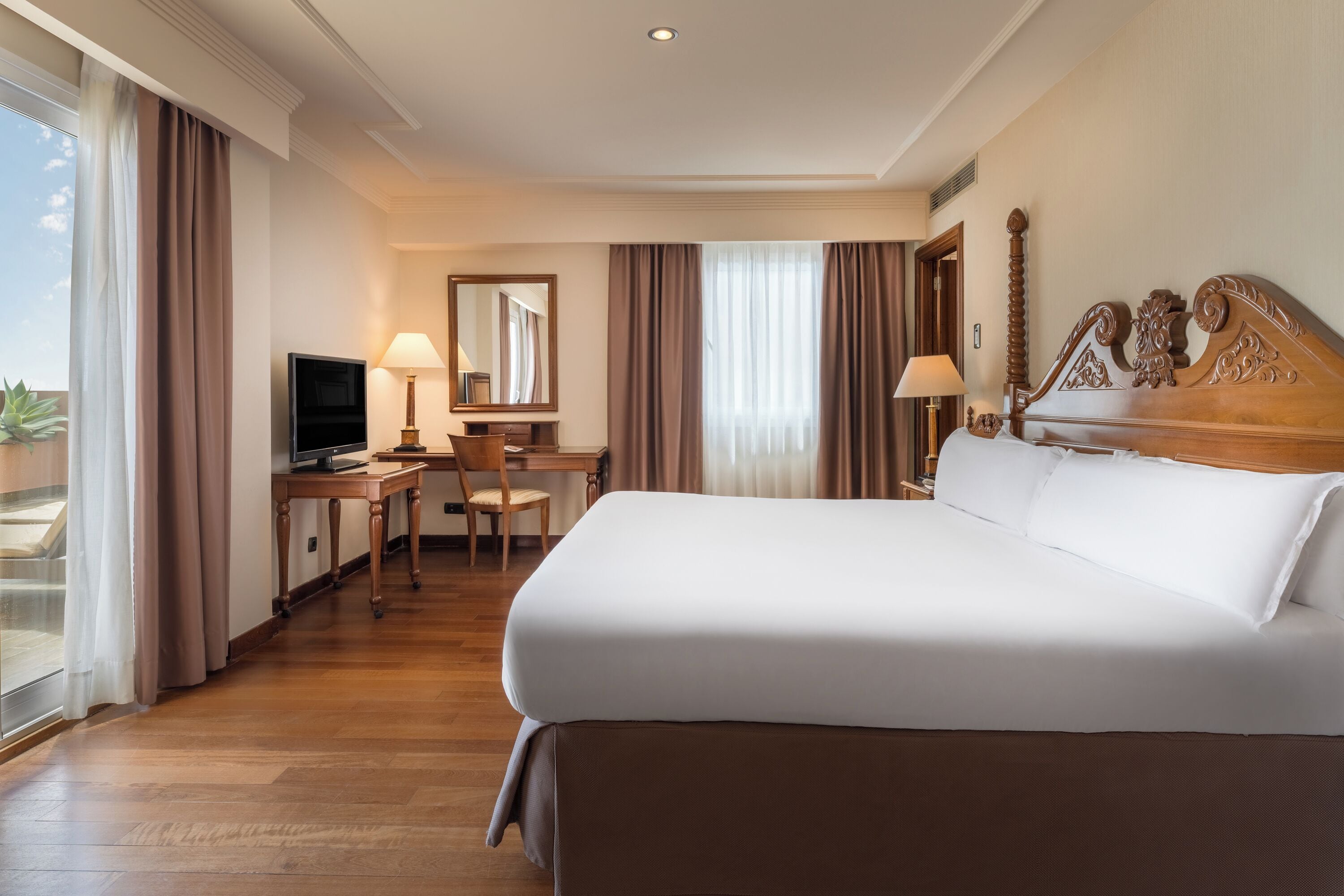 A hotel room with a large bed, a desk with a chair, a television, and a window with curtains.