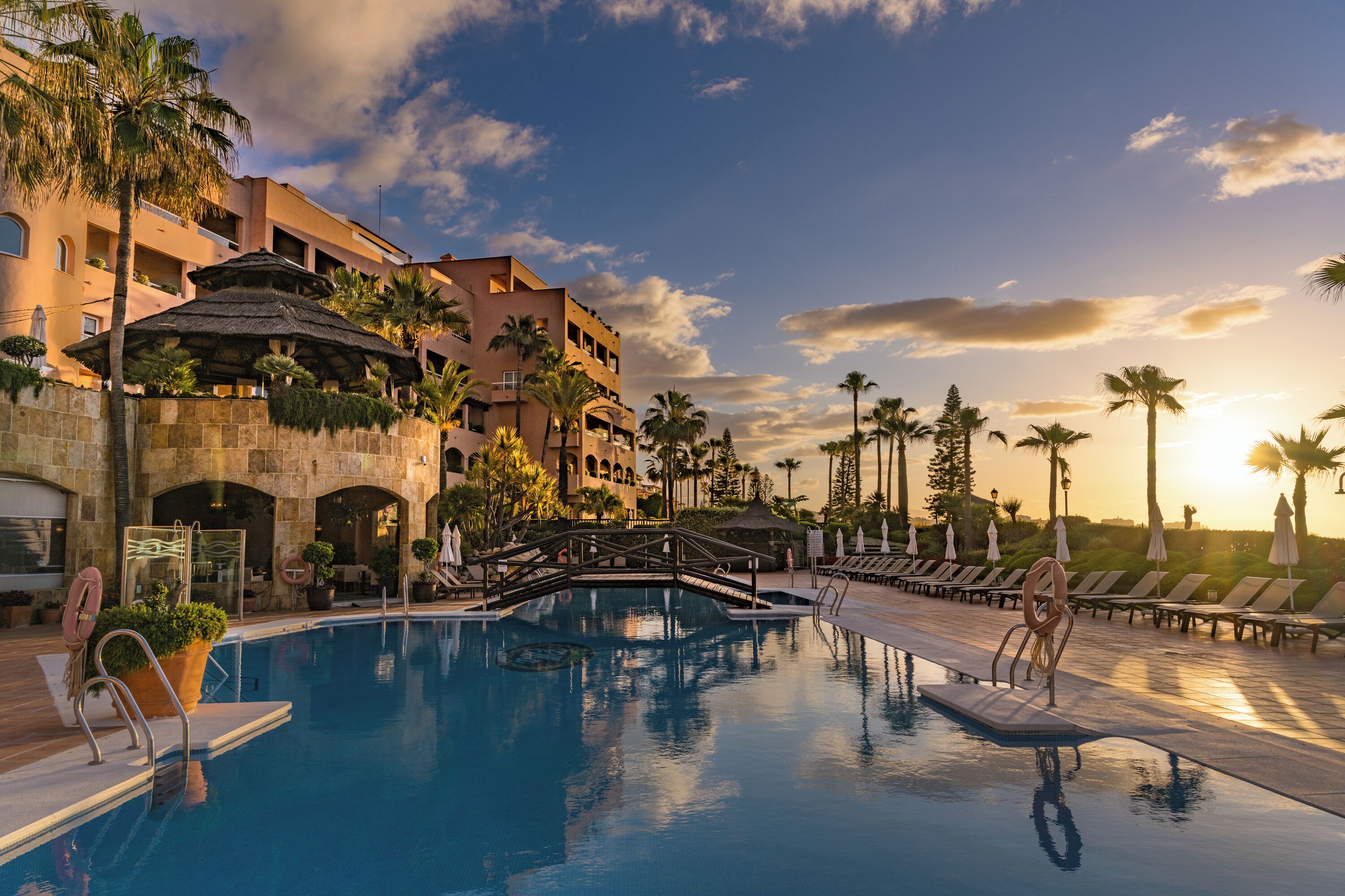 A pool area with a bridge, lounge chairs, and palm trees.