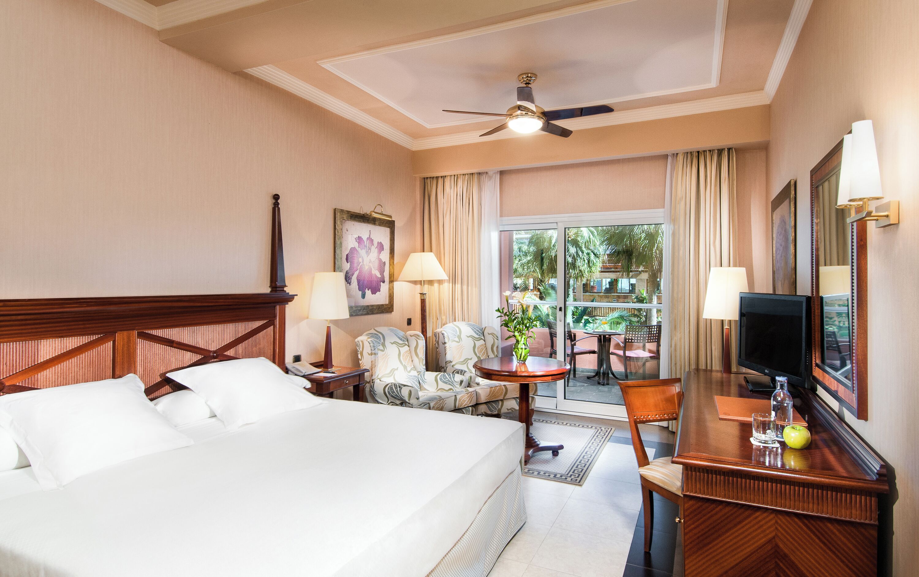 Quarto de hotel com cama, escrivaninha, cadeira, televisão e ventoinha no teto.