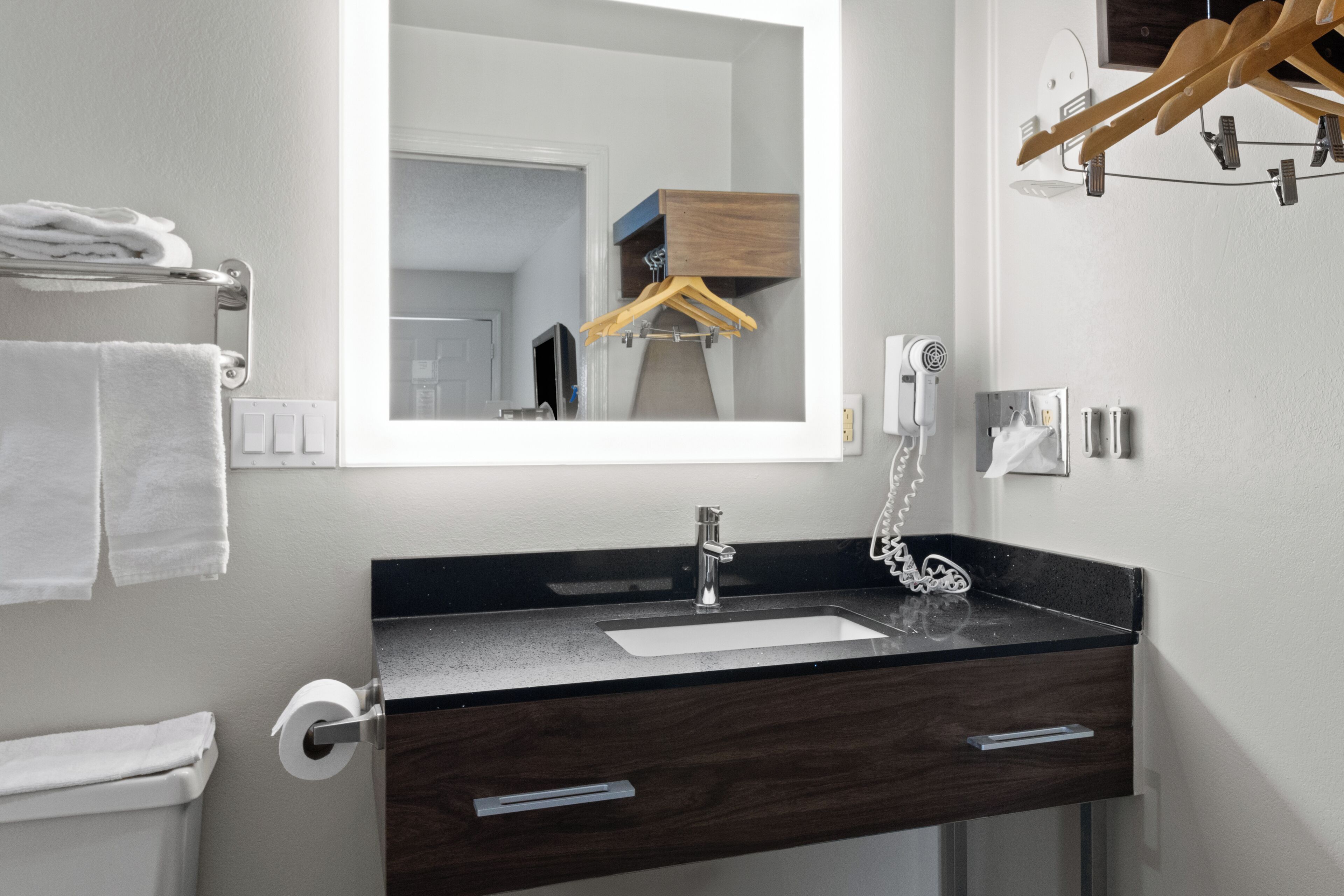 Un baño moderno con tocador de madera oscura, un espejo grande, un lavabo blanco y un perchero con toallas dobladas.