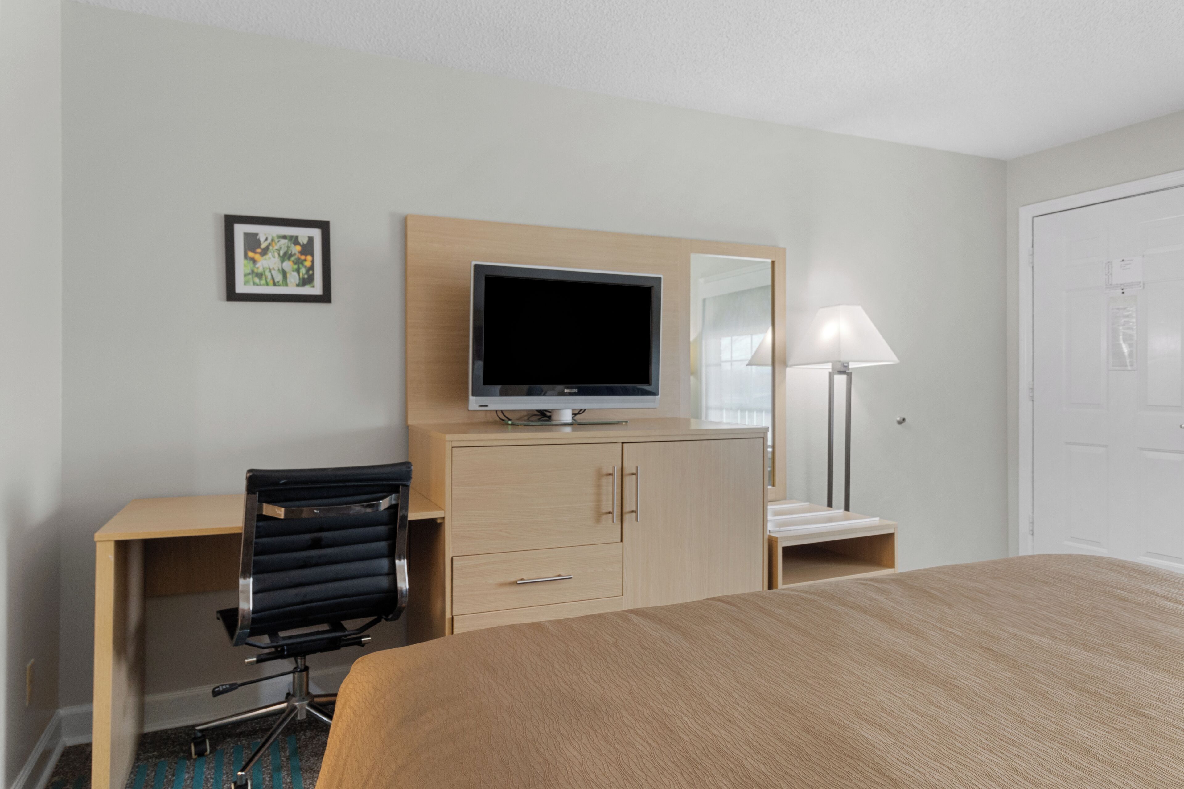 A hotel room with a bed, a desk with a chair, a television, a lamp, and a wardrobe.