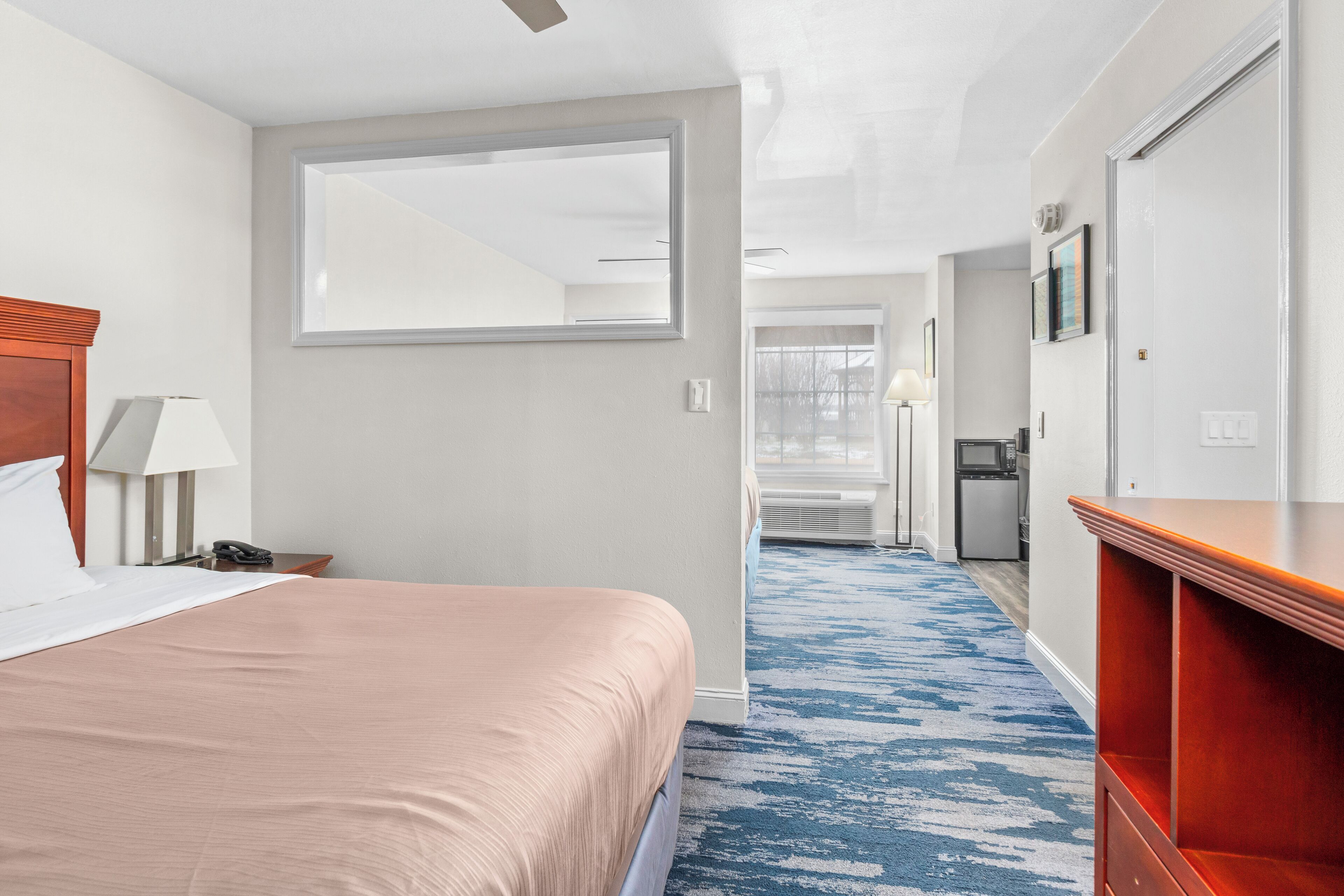 Habitación de hotel con cama, escritorio, silla, lámpara, espejo, armario y baño visible a través de una puerta abierta.