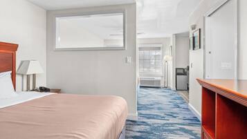 Habitación de hotel con cama, escritorio, silla, lámpara, espejo, armario y baño visible a través de una puerta abierta.