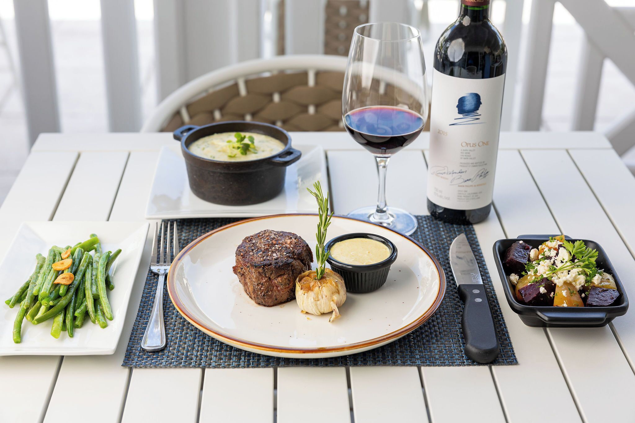 A dining table with a steak, green beans, a bowl of soup, a glass of red wine, and a bottle of wine.