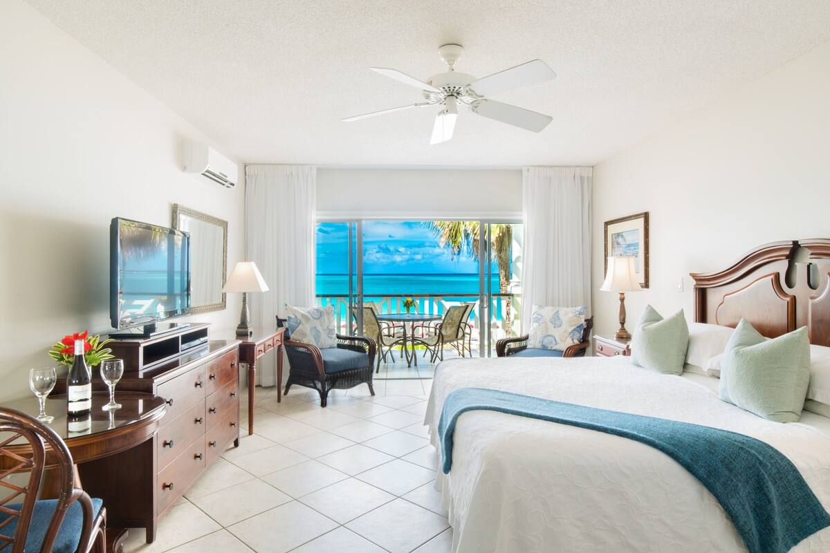 Een hotelkamer met een bed, een bureau met een televisie, een stoel, een tafel met een fles en glazen, en een balkon met uitzicht op zee.