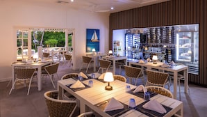 Un restaurant avec des tables blanches et des chaises en osier, un coin bar avec des bouteilles et des verres, et une vue sur la verdure à l’extérieur.