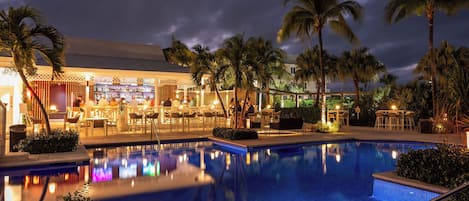 Zona de piscina con bar y asientos, rodeada de palmeras y un edificio con terraza cubierta.