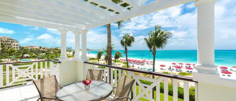 A covered outdoor area with a table and chairs, overlooking a beach with umbrellas and palm trees.