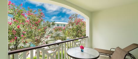 A balcony with a round table and two chairs, a white railing, and a view of pink flowers and a building.