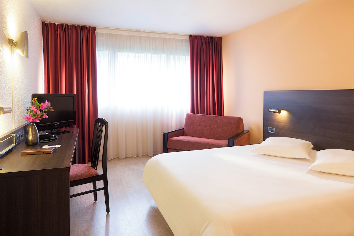 A hotel room with a bed, a desk with a TV, a chair, and a window with curtains.