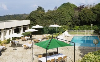 Zona de piscina al aire libre con sillones de descanso y sombrillas, rodeada de setos y un edificio.