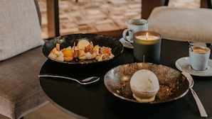 Una mesa de comedor con platos de comida, una vela y una taza de café en un salón moderno con una estantería y asientos cómodos.