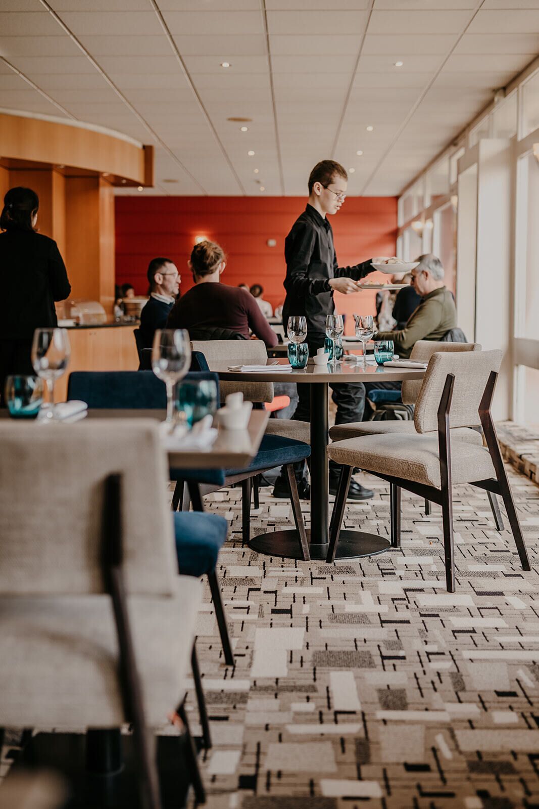 A modern dining area with tables and chairs, a waiter serving a customer, and a patterned floor.