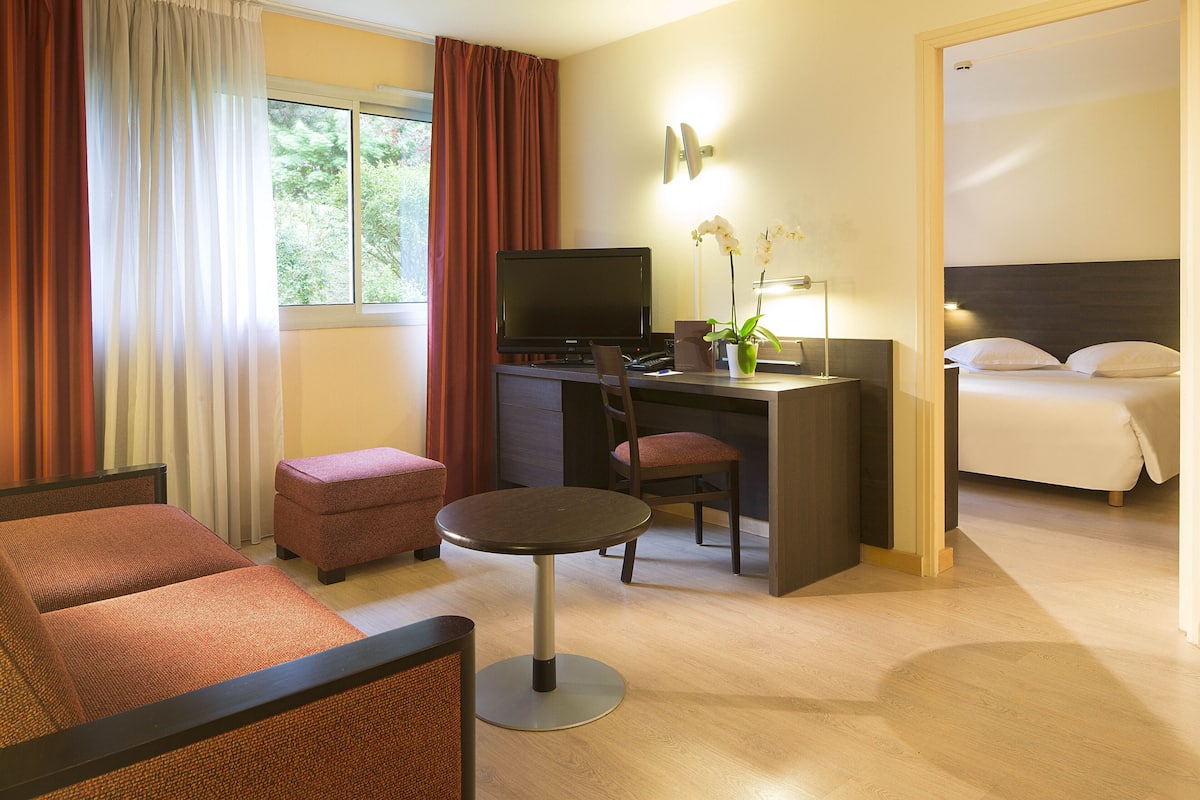 A hotel room with a bed, a desk with a computer, a chair, a small table, and a window with curtains.