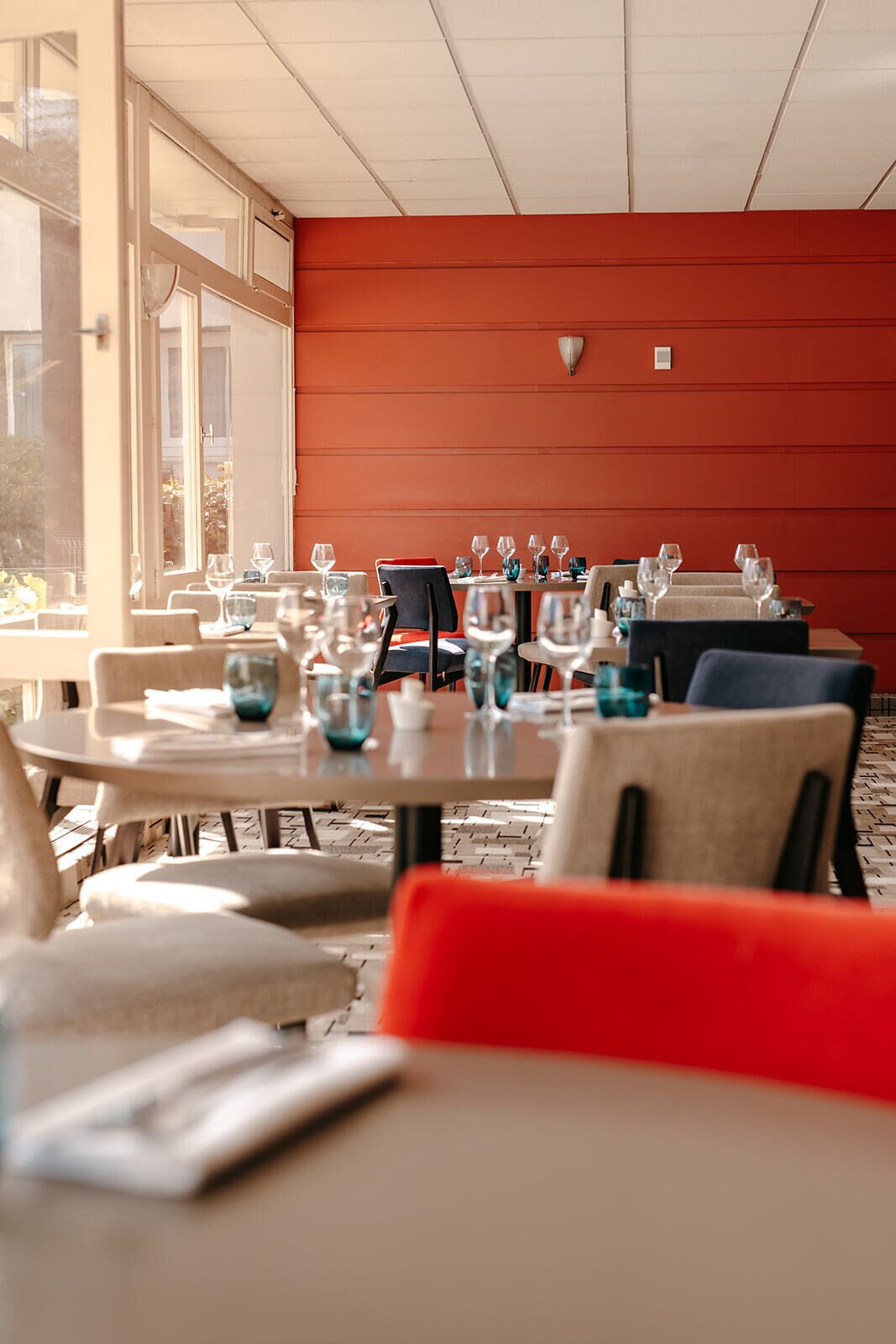 A restaurant with tables set for dining, large windows, and a red wall.