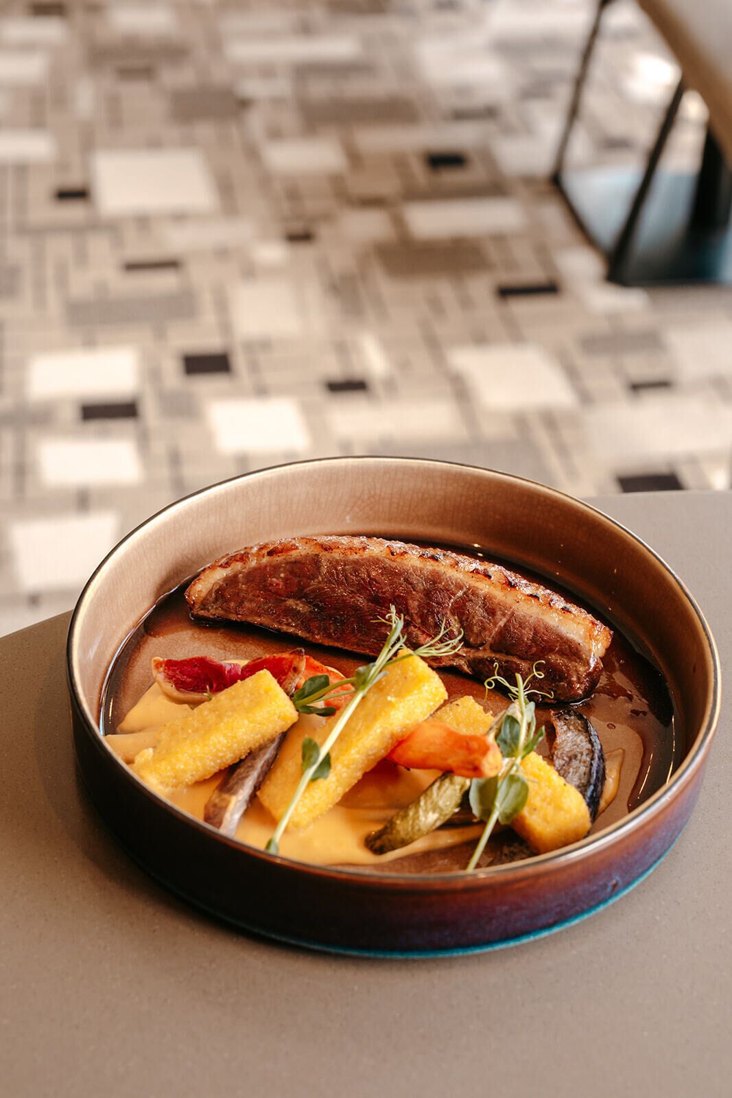 A bowl of food with a piece of meat and vegetables on a table.