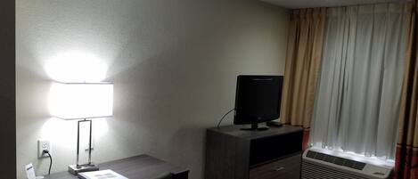 A hotel room with a desk, a flat-screen TV, a lamp, a heater, and a window with curtains.