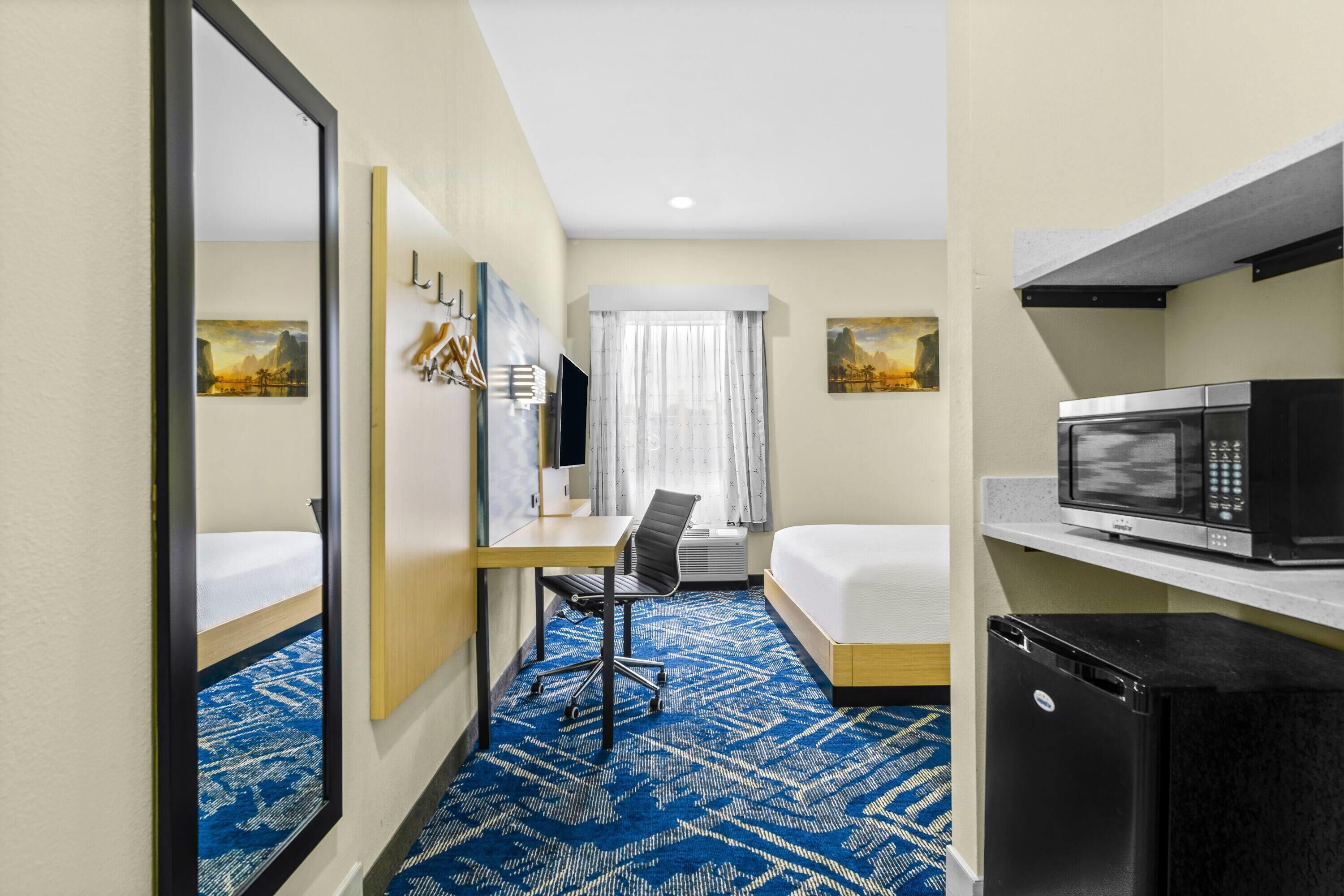 A hotel room with a bed, desk, chair, microwave, refrigerator, and a large mirror.