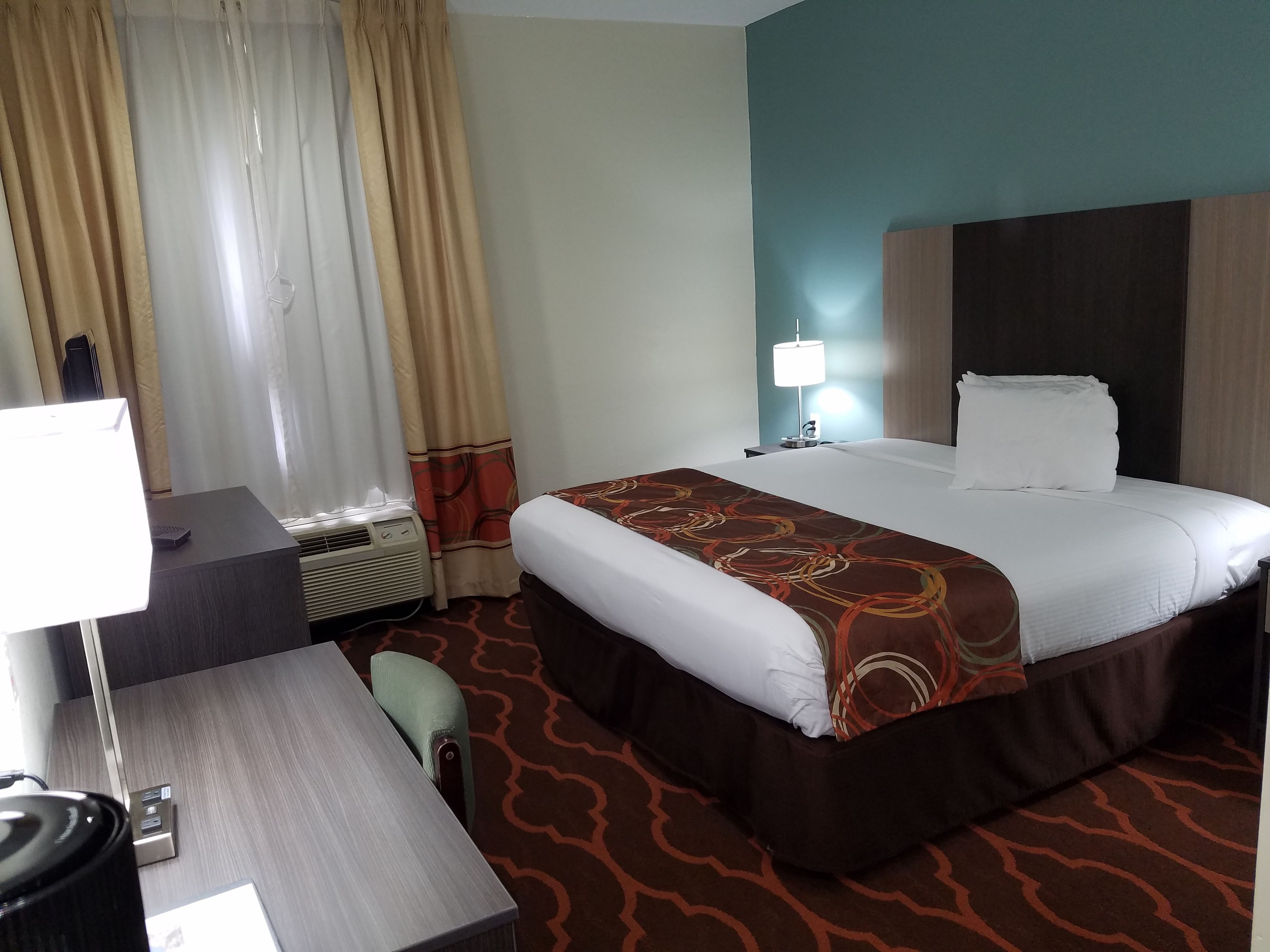 A hotel room with a bed, bedside table, lamp, air conditioning unit, and a desk with a computer.