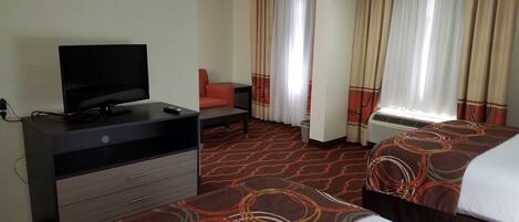 Habitación de hotel con una televisión sobre un tocador, dos camas con cubrecamas estampados, una silla y un ventanal con cortinas.