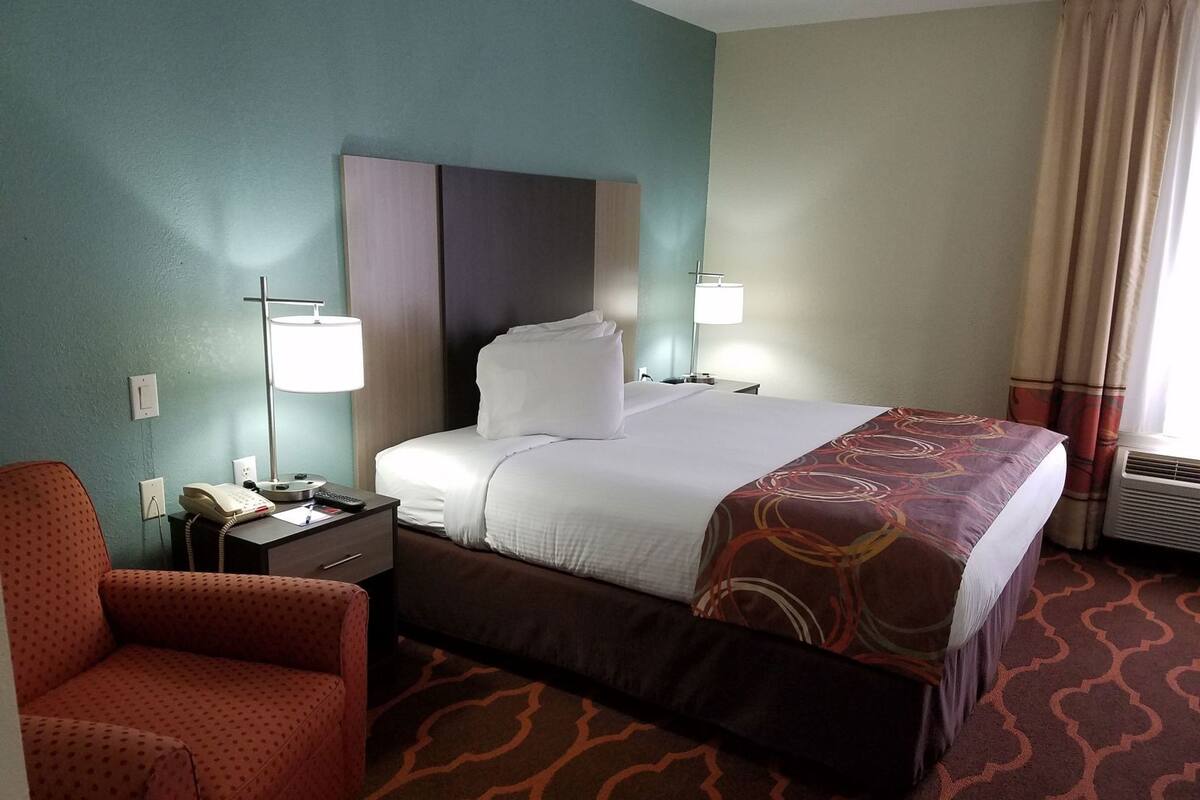 A hotel room with a bed, a desk with a phone, a chair, a lamp, and a window with curtains.