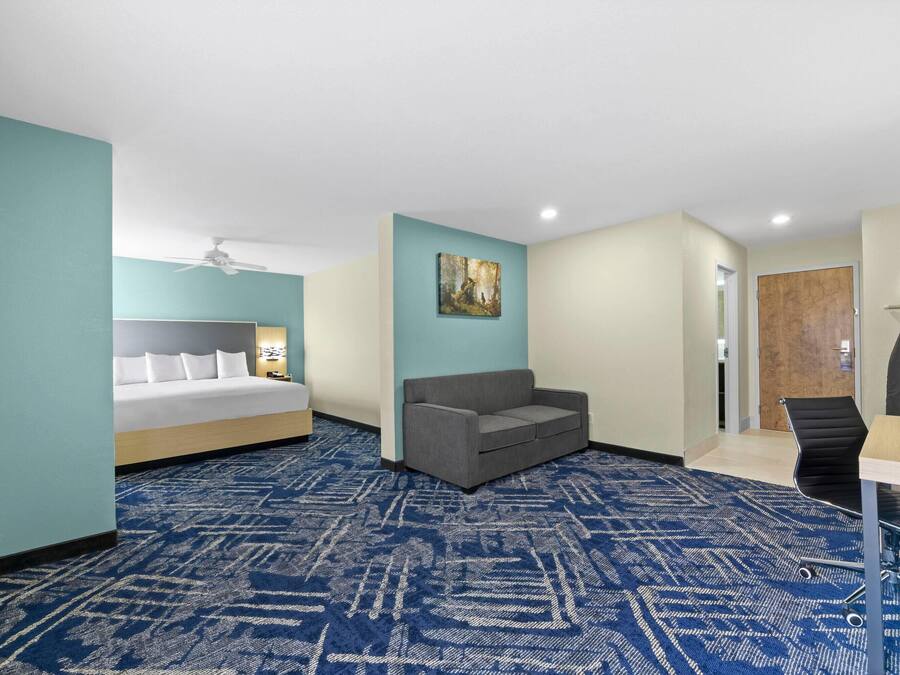Habitación de hotel con cama, sofá gris, escritorio con silla y un cuadro en la pared.