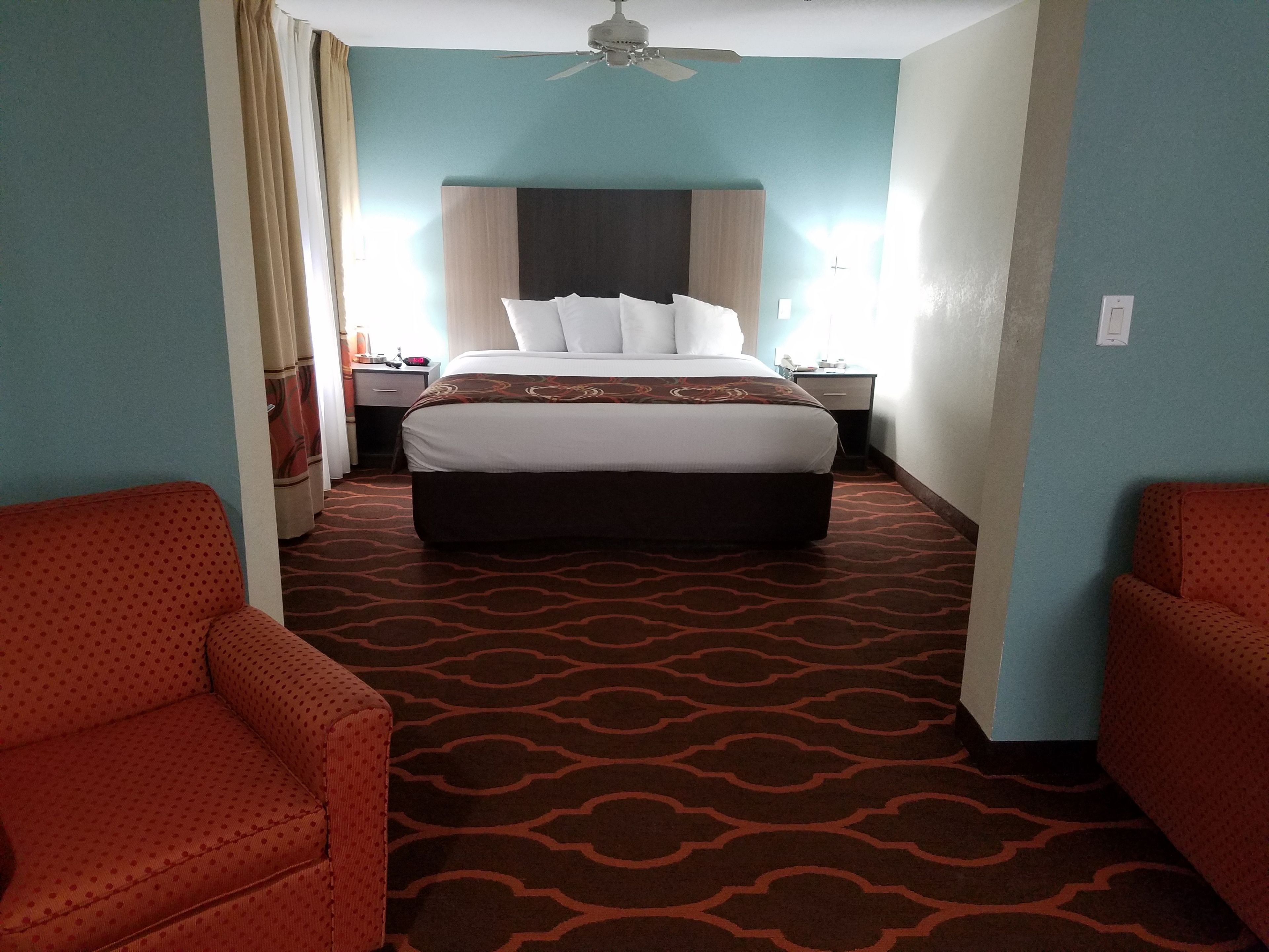 A hotel room with a large bed, two red armchairs, a ceiling fan, and a patterned carpet.