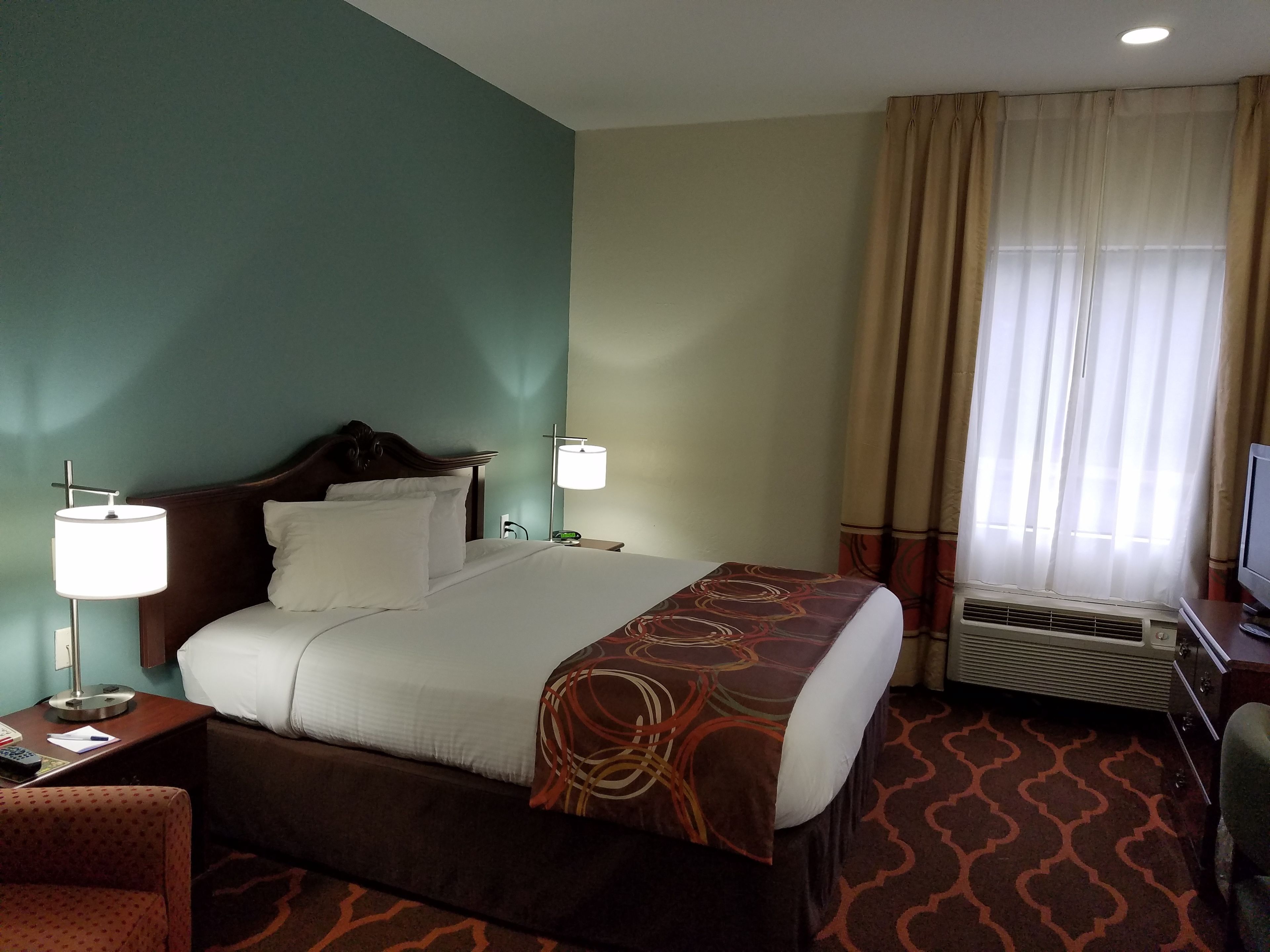 A hotel room with a large bed, two bedside tables with lamps, a television, and an air conditioning unit.