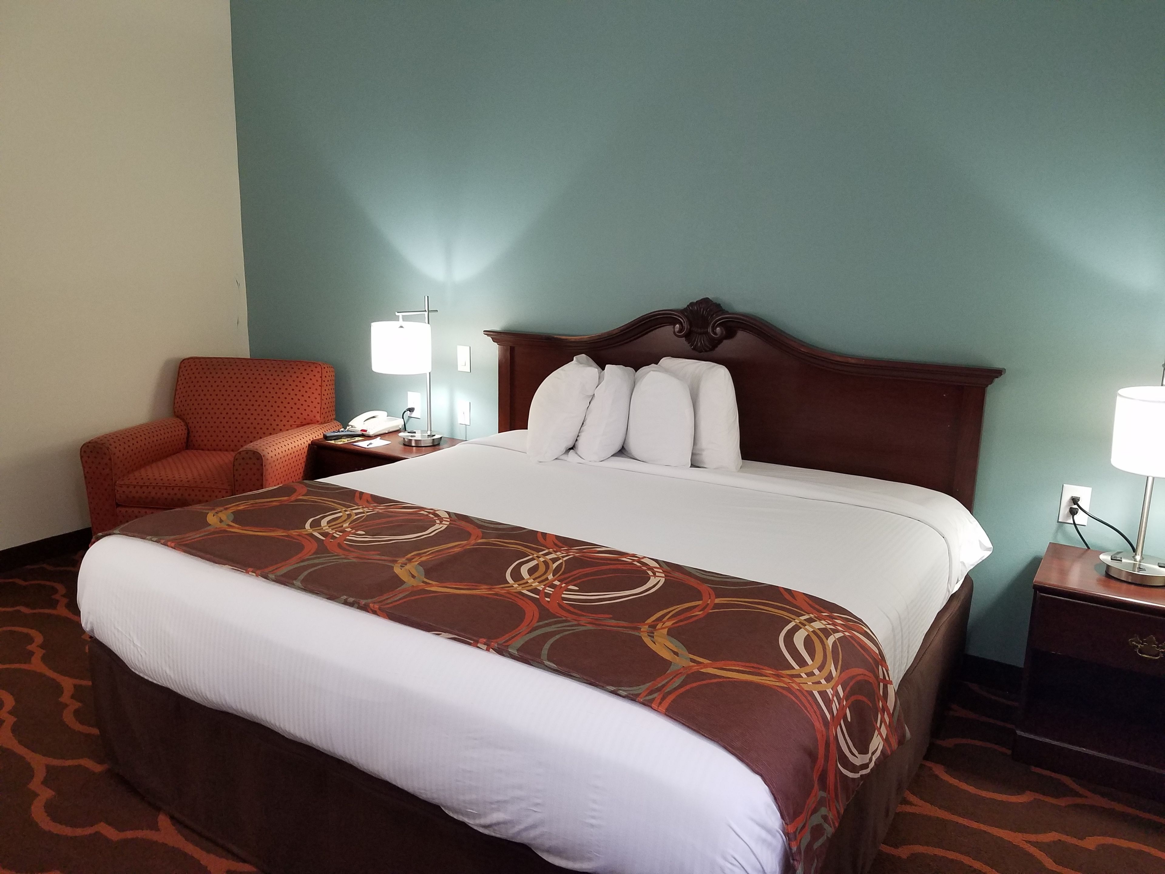 Habitación de hotel con una cama grande, una silla, una mesita de noche con lámpara y un enchufe de pared.