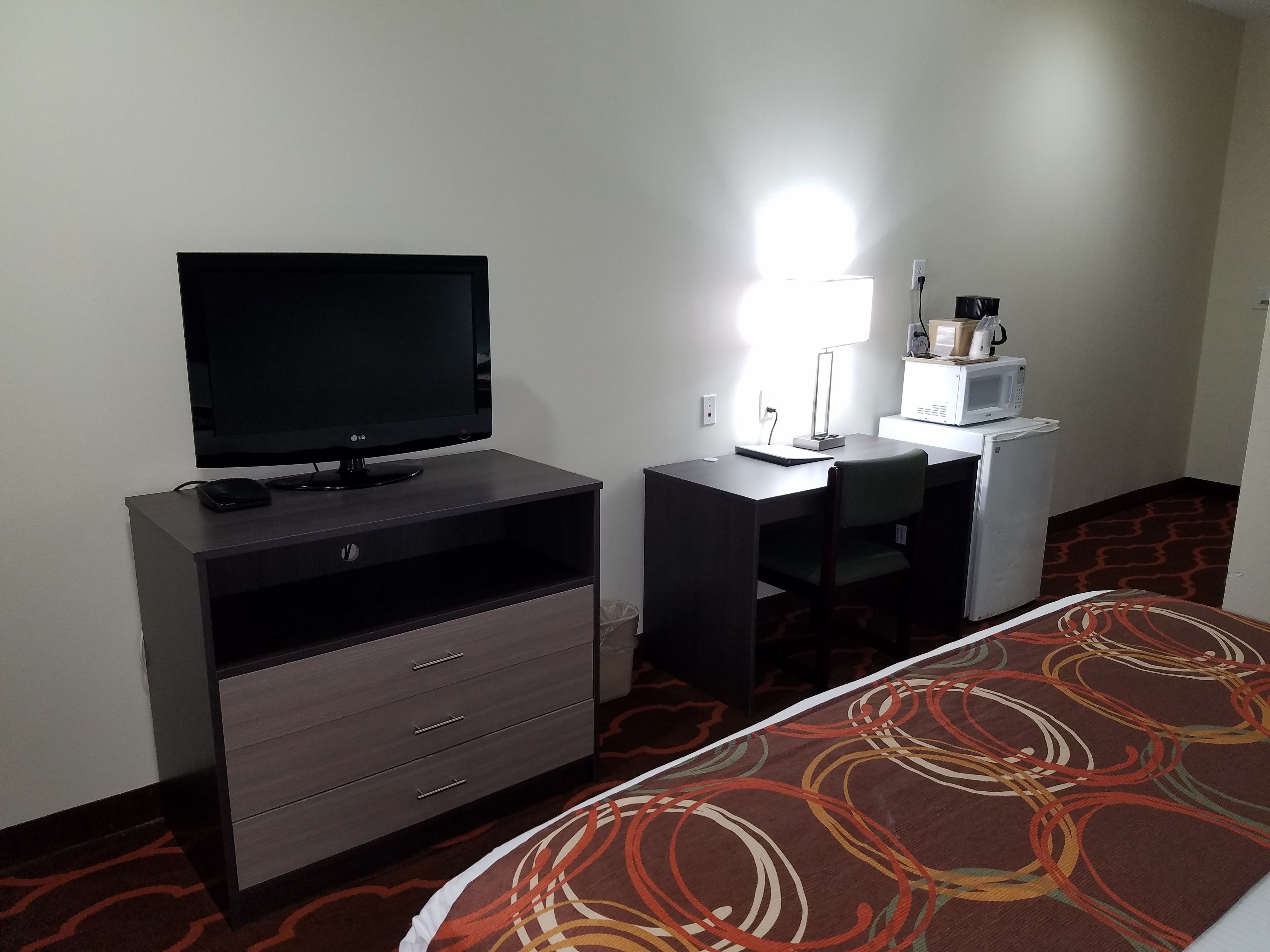 A hotel room with a TV on a wooden cabinet, a desk with a lamp, a microwave, and a bed with a patterned bedspread.