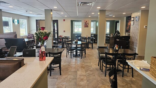 A restaurant with multiple tables and chairs, a counter with a cash register, and a wall-mounted menu board. - Baymont by Wyndham Grovetown Augusta (Grovetown)